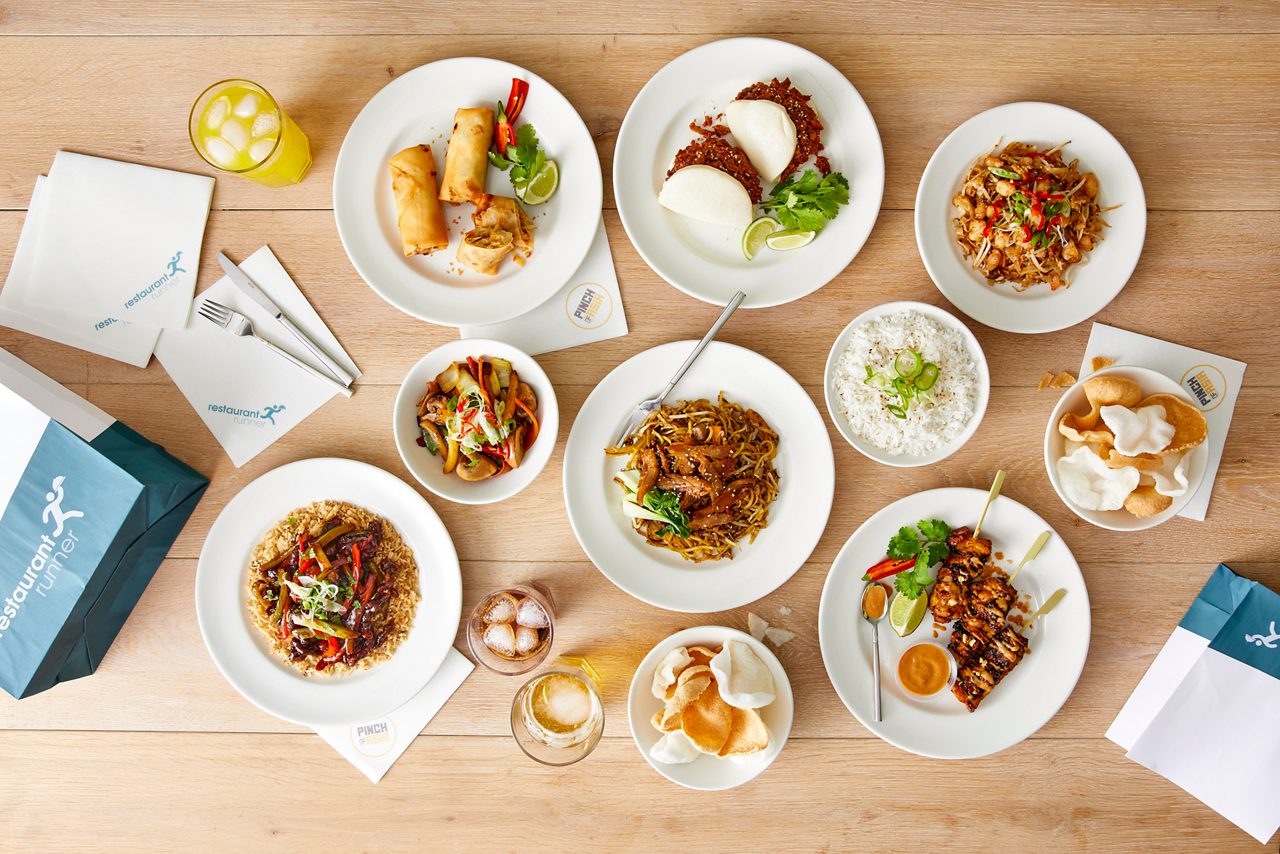 Assorted Asian dishes sit arranged on a wooden table; spring rolls, noodles, rice, satay skewers, stir-fries, and prawn crackers accompany drinks and delivery bags. Text: restaurant runner; PINCH.